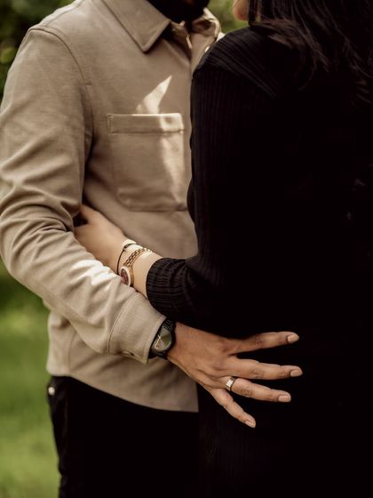 A close-up in warm monochrome, focusing on the couple's embrace and their hands on the baby bump. The sunlight filtering through the trees adds a beautiful, gentle light.