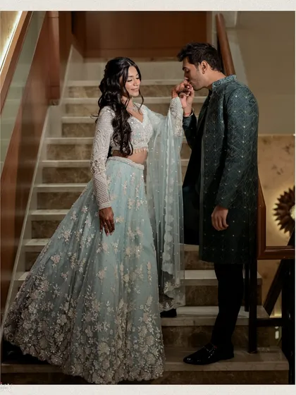 A classic and romantic pose on a grand staircase. The groom gently kisses the bride's hand, a timeless gesture of love, while her flowing light blue lehenga drapes beautifully down the steps.