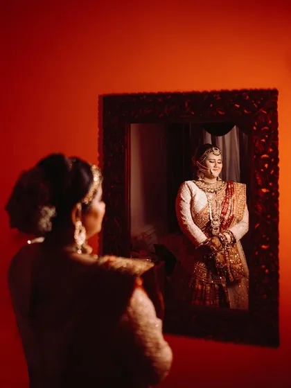 A beautiful mirror portrait of the bride. Her reflection is perfectly captured, showcasing her full bridal attire and serene smile. The red background adds a touch of passion and drama.
