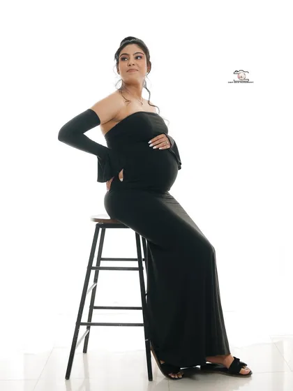 A chic and modern solo portrait. The mother-to-be is seated on a stool against a clean white background, wearing a sleek black off-the-shoulder dress that elegantly shows off her silhouette.