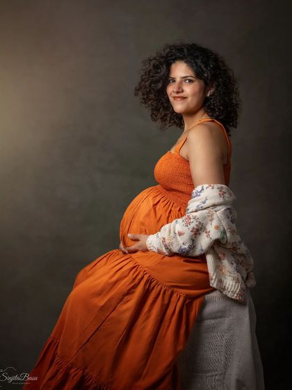 A duplicate of ID 12, this is a warm and gentle studio portrait of a mother-to-be in a comfortable orange dress, her curly hair framing a soft smile. The simple, classic backdrop keeps the focus entirely on her serene expression.