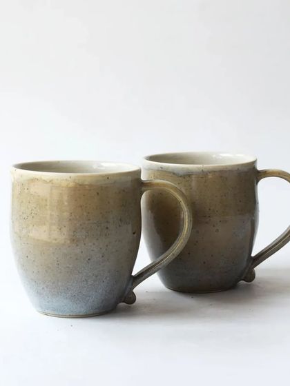 A pair of our classic mugs in a beautiful gradient glaze that shifts from earthy brown to a soft blue. The subtle details, like the small nub at the base of the handle, showcase their handmade quality.
