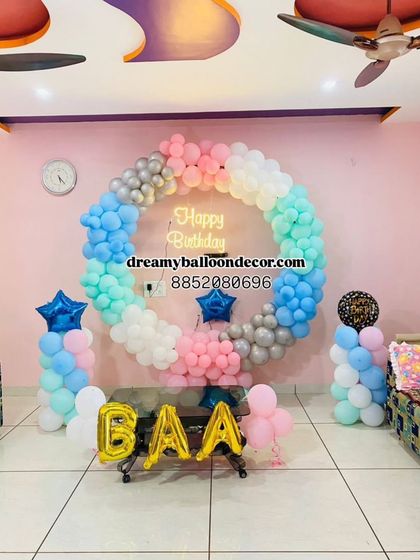 A beautiful pastel-themed birthday decoration for a home setting. The balloon ring features shades of blue, pink, and mint green, with a 'Happy Birthday' neon sign and custom foil letter balloons.