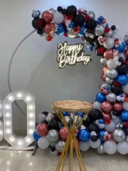 A modern and sleek setup for a 30th birthday. The silver ring frame is partially decorated with a balloon garland in black, silver, red, and blue, paired with a neon sign and marquee numbers.