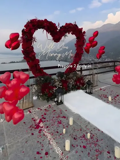 Leveraging a stunning natural view, this terrace proposal features a large heart-shaped arch made of deep red flowers. A white aisle runner, scattered petals, LED candles, and heart-shaped balloons create a clear path for a memorable walk-up to the big question.