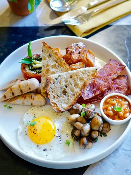 A classic full breakfast, perfectly plated and ready to enjoy. This hearty meal includes a sunny-side-up organic egg, sausages, bacon, baked beans, sautéed mushrooms, and sourdough toast, showcasing our commitment to quality ingredients for your morning meal.