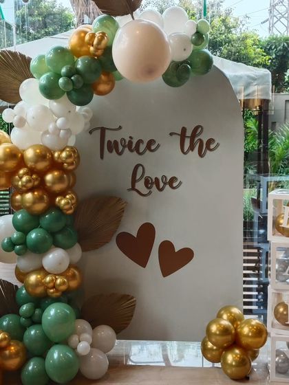 A beautiful setup for a baby shower celebrating twins, with the message 'Twice the Love'. The decor uses an arched backdrop and a sophisticated balloon garland in sage green, white, and chrome gold.