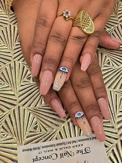 Long, coffin-shaped nude nails with a trendy twist. The accent nails feature hand-painted evil eye designs, with a silver glitter base at the cuticle for extra flair.