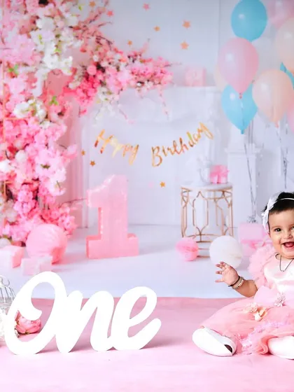 A happy one-year-old girl in her pink party dress, surrounded by a beautiful floral and balloon arrangement for her birthday photoshoot.