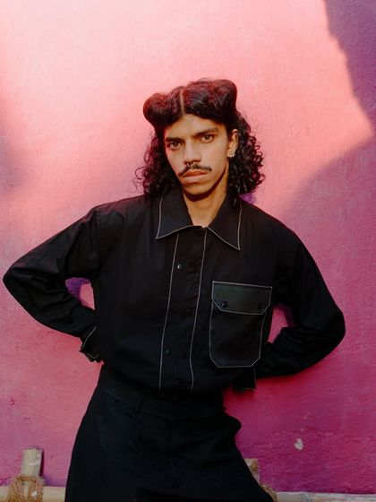 A bold, colorful shot from a menswear editorial in Goa. The sharp contrast between the all-black outfit with white piping and the vibrant pink wall creates a visually arresting image.
