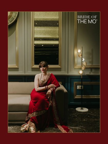 A stylish, editorial portrait titled "BRIDE OF THE MO'." The bride is seated on a sofa in a modern, well-lit room in Jammu, her red saree creating a bold statement.