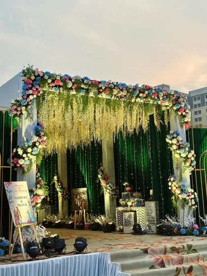 A beautiful reception stage with an emerald green velvet backdrop and hanging wisteria flowers. The setup is adorned with pastel floral arrangements and modern gold-accented furniture.