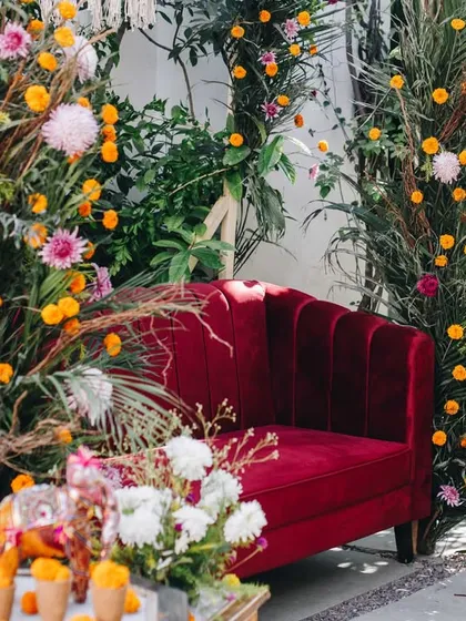 A vibrant and lush photo corner for a Mehendi, featuring a plush velvet sofa surrounded by a profusion of colorful flowers and greenery.