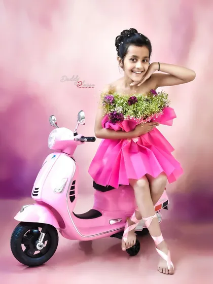 A fun and stylish shot of a ten year old girl in a unique pink dress made of flowers, posing on a matching pink toy scooter. This shows a creative concept for older kids' photoshoots.