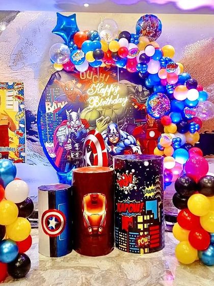 A large-scale Avengers setup with a custom backdrop, balloon garland, and themed pedestals for cake and gifts.