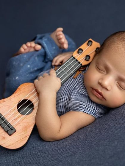 A tiny musician in the making. This baby boy is dressed in a stylish blue outfit and is holding a miniature ukulele, looking perfectly peaceful and dreaming of sweet melodies.