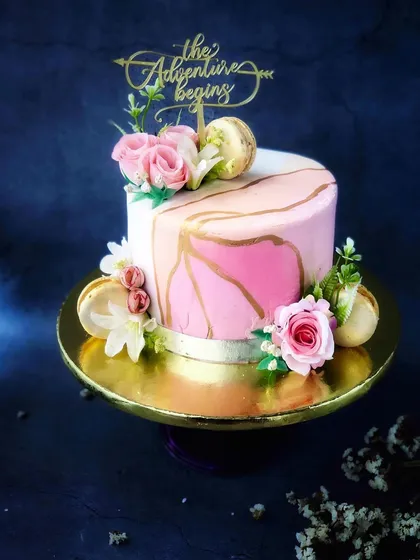 A chic and modern engagement cake with a pink marble effect, gold accents, and fresh flowers, topped with "The Adventure Begins."