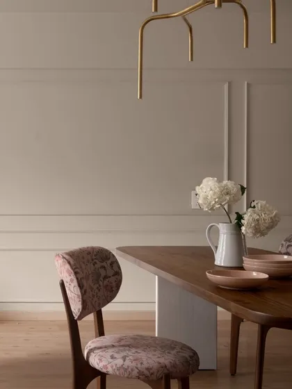 The dining chairs in "Apartment 222" are upholstered in a delicate paisley fabric, adding a touch of pattern to the otherwise minimalist space. The warm wood of the table and the soft pink of the bowls create a welcoming tablescape.