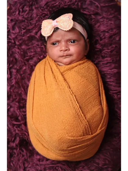 A classic "potato sack" pose, with the newborn swaddled snugly in a mustard-colored wrap. This simple portrait on a deep purple background highlights the baby's adorable, alert expression.
