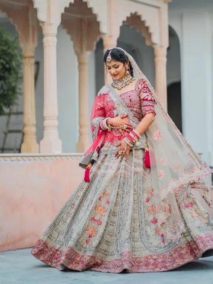 A full-length portrait of the bride Ashri, showcasing her magnificent multi-colored floral lehenga from Kalki Fashion. The architectural background adds a touch of royal elegance.