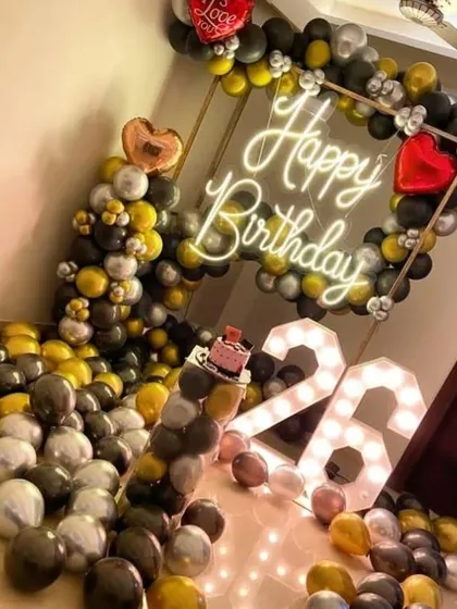 A cool and stylish 26th birthday setup. The room is filled with black, yellow, and silver balloons, with a neon sign, light-up numbers, and a small cake ready for celebration.