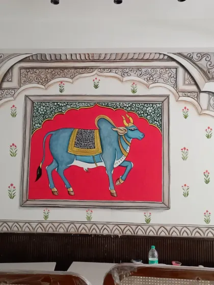 A classic Pichwai style painting, this mural brings serenity and tradition into the home. Featuring a beautifully adorned cow against a vibrant red backdrop, framed by intricate Rajasthani architectural motifs, it is a piece of heritage art that creates a divine atmosphere.