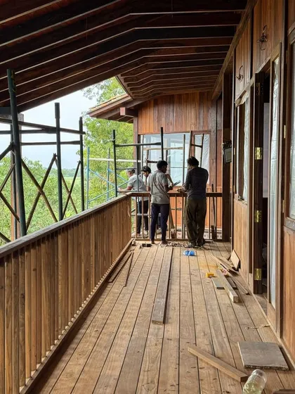 Workers install window frames on the upper-level balcony. The extensive use of wood for the floors, walls, and railings creates a warm and cohesive architectural language throughout the project.