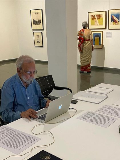 The artist working on his laptop at the library table, with his wife Nilima Sheikh viewing the hung works in the background.