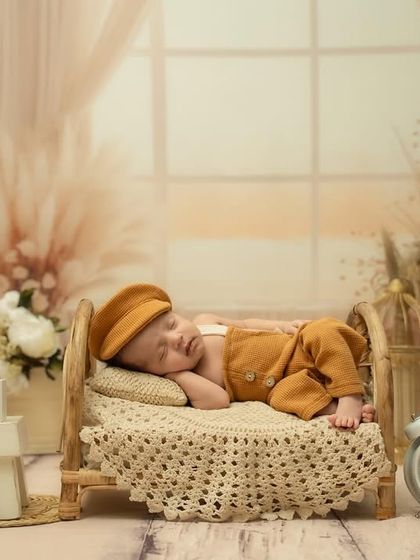 A newborn in a vintage-style outfit and cap sleeps on a miniature wooden bed in a beautifully arranged rustic studio setup.