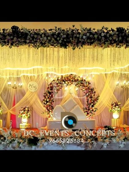 This wide stage design for a reception features a beautiful floral arch, warm curtain drapes, and a ceiling of hanging tassels and fairy lights for a layered, luxurious effect.