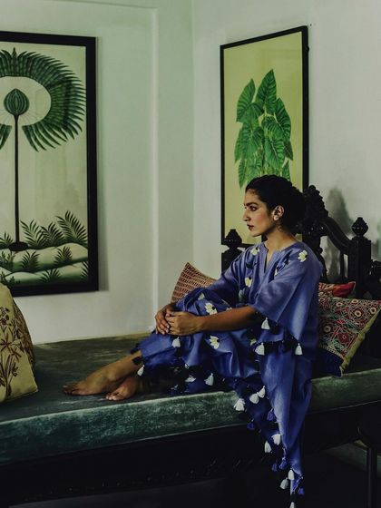 A pensive portrait for Baavli, with the model seated in a classic, art-filled room. The composition feels like a painting, capturing a quiet moment of reflection while showcasing the elegant drape of the tassel-hemmed kaftan.