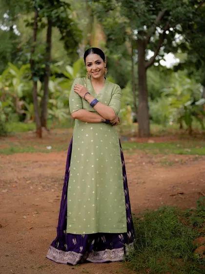 A full-length portrait of actress Anupama Gowda in a traditional green and purple outfit. The natural setting and her confident pose create a strong and elegant image.