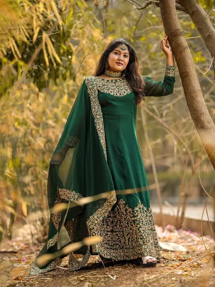 A regal full-length portrait of a woman in a forest green Anarkali with heavy gold embroidery, leaning against a tree.