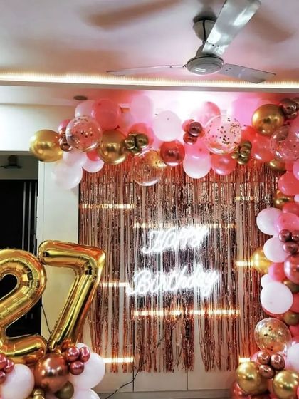 A chic 27th birthday decoration with a rose gold theme. The balloon arch and fringe backdrop are complemented by a neon sign and large number balloons.