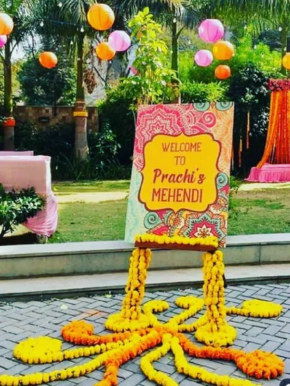 We created this vibrant outdoor Mehendi setup on a spacious lawn, designed to be both welcoming and picturesque. The personalized "Welcome to Prachi's Mehendi" sign, set on a wooden easel with marigold garlands, greets guests as they arrive. Overhead, colorful paper lanterns in pink and orange add a festive touch, complementing the decorated seating area visible in the background.