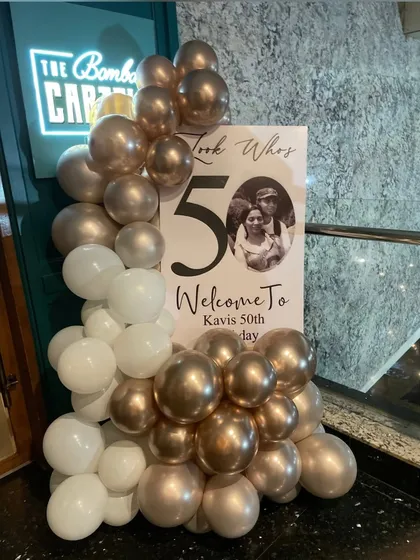 A personalized welcome sign for a 50th birthday party. This U-shaped board features a custom photo and message, framed by an elegant white and champagne gold organic balloon garland to greet guests in style.