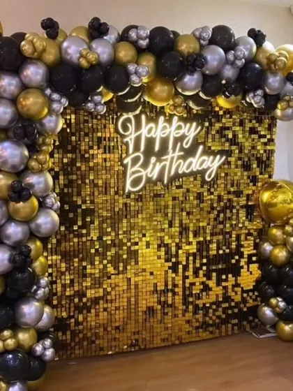 A close-up of a glamorous golden sequin wall with a 'Happy Birthday' neon sign, framed by black, silver, and gold balloons.