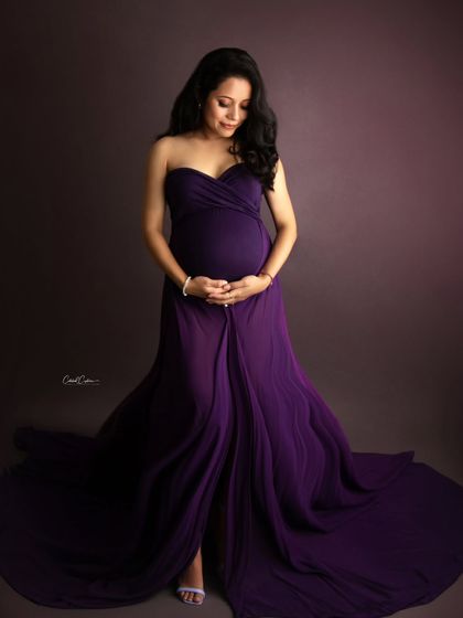 A classic and elegant studio portrait. The mom-to-be looks down, cradling her bump in a simple yet stunning deep purple gown.