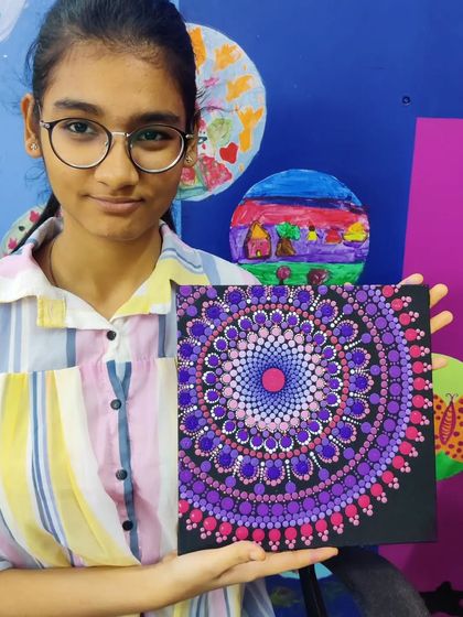 A young adult proudly holds up her completed dot mandala painting. The design, featuring shades of purple and pink with a gradient effect, is a beautiful example of the complex patterns we explore in my workshops.