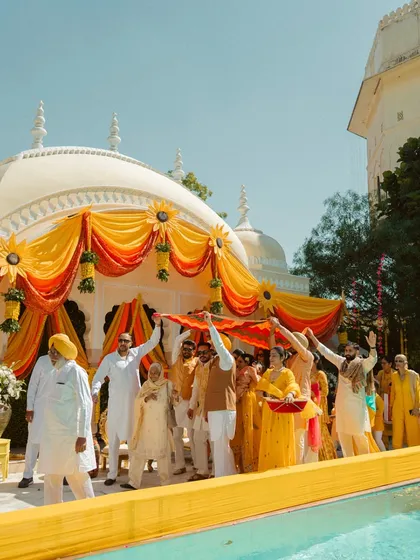 A wide shot of the Haldi ceremony entrance, showing family and friends welcoming the couple under a vibrant yellow and orange canopy.