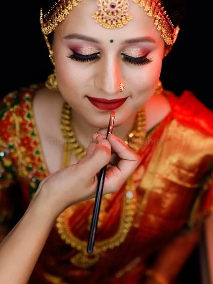 A classic muhurtham look featuring a bright red lip and defined eyes. The traditional matha patti and heavy gold jewellery are perfectly balanced with the makeup, and the bridal braid with fresh flowers is a timeless choice.