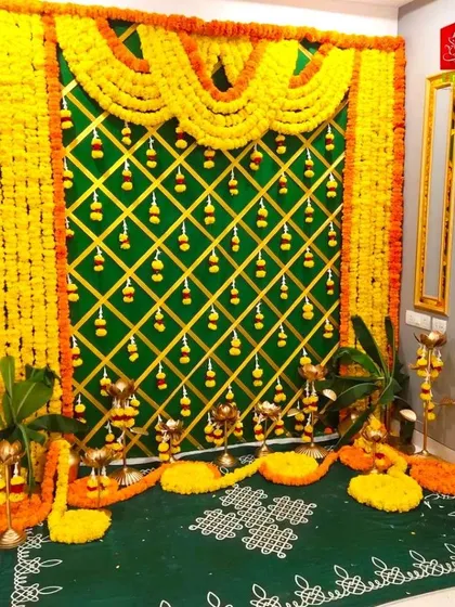 A traditional baby shower setup with a marigold and banana leaf backdrop. The decor includes a green floor mat with rangoli and traditional lamps.