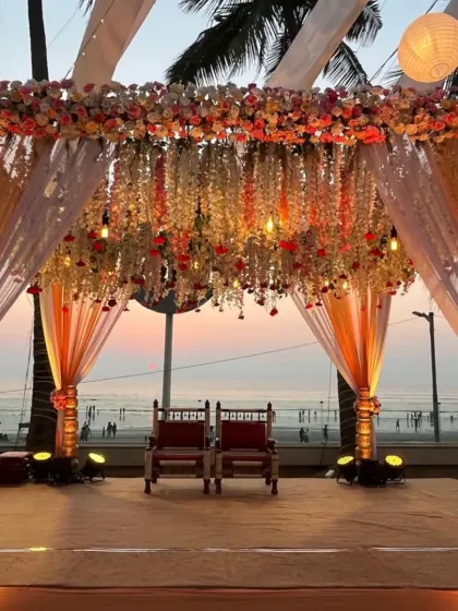 This symmetrical shot captures the beauty of a beach mandap at dusk. The structure, with its intricate floral ceiling and traditional pillars, is perfectly illuminated against the fading light of the sunset.