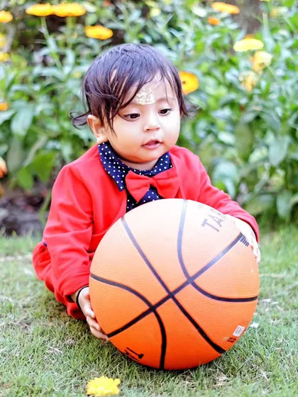 A young boy in a red suit jacket holds a basketball during a sports-themed outdoor photoshoot in a garden.