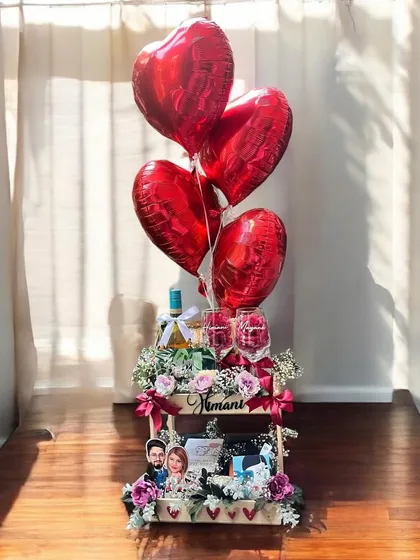 I designed this romantic anniversary hamper to be a showstopper, featuring a large wooden crate filled with flowers, personalized photo frames, wine, and elegant glasses, all topped with heart-shaped red foil balloons.