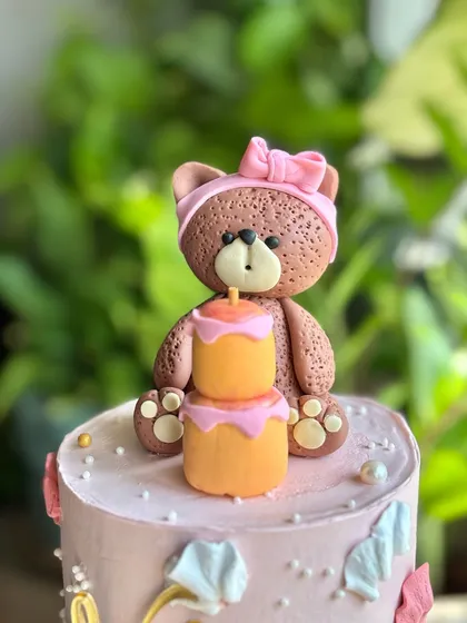 A detailed shot of the adorable fondant teddy bear topper, complete with a pink bow and a tiny two-tier cake with a candle.