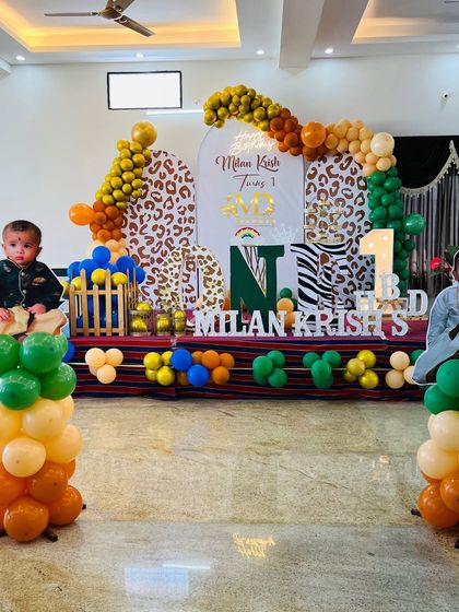 A full stage setup for a jungle-themed first birthday with animal print backdrops. The decor includes life-size cutouts of the birthday boy, a colorful balloon arch, and his name in large letters.