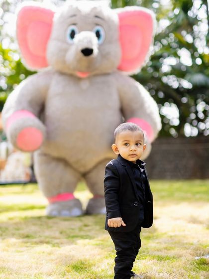 The birthday boy looking sharp in his little suit, standing in front of a large elephant mascot at his safari-themed party.