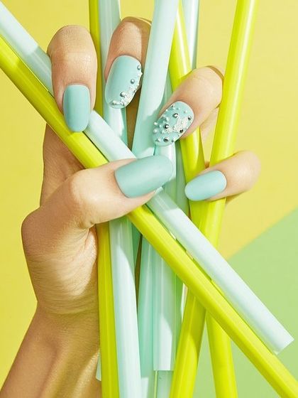 A close-up of a hand with a fresh, mint-green manicure holding matching straws. The bright, color-blocked background makes this a fun and summery shot for a nail polish brand.