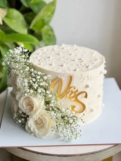 A Pinterest-worthy single-tier cake perfect for a roka or intimate wedding. It features a cascade of fresh white roses and baby's breath, a custom gold initial topper, and delicate pearl details.